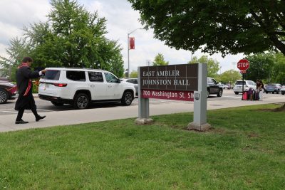 A graduating Hokie walks past the sign for East Ambler Johnston Hall, dressed for a Commencement ceremony. Many cars park along the street for move-out behind him, and two students stand near a stop sign with luggage waiting to be picked up after moving out of their residence hall. 